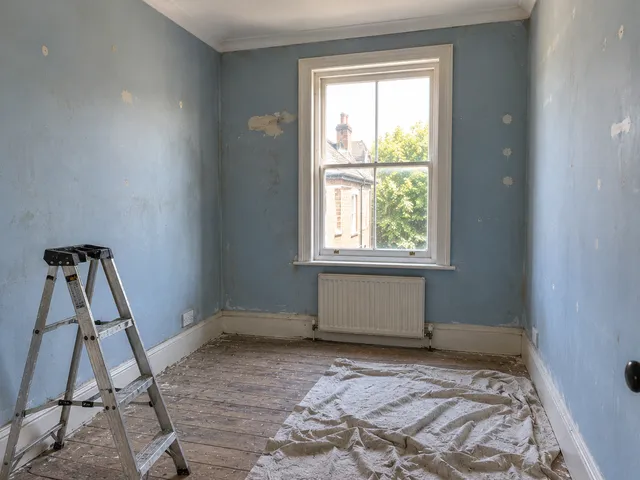 A small bedroom with patchy, peeling blue walls, a stepladder and dust sheets.