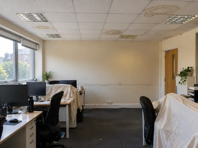 An office with water-stained ceiling tiles, marked walls and desks under dust sheets.