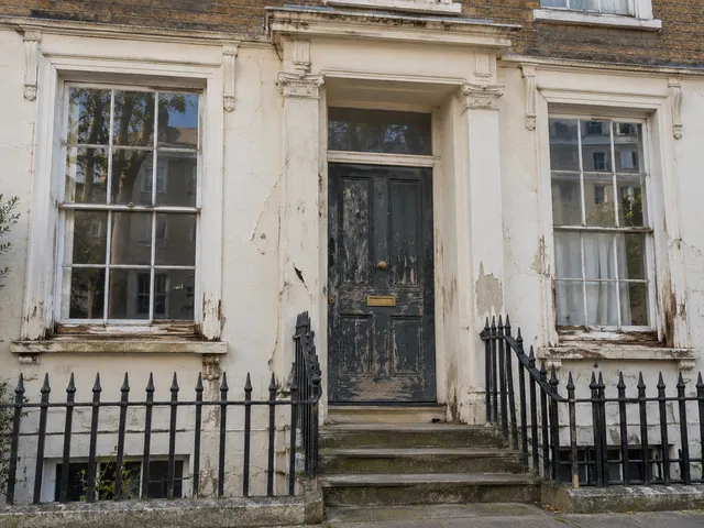 A townhouse front with flaking sash windows, cracked render and a worn front door.