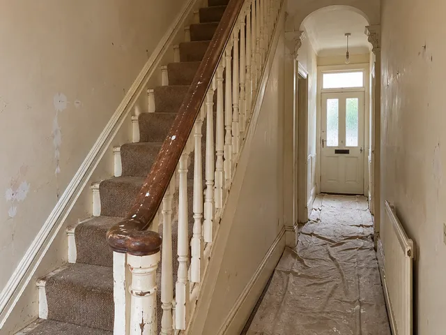 A period hallway with stained, patchy walls, chipped spindles and dust sheets on the floor.